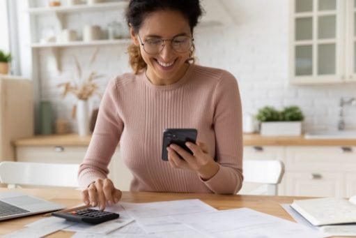 A smiling woman sits at her kitchen table looking at her phone. A laptop, papers, and calculator are on the tabletop.