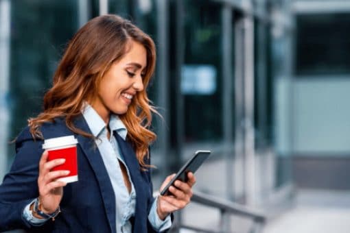 Smiling businesswoman viewing mobile phone and enjoying coffee break in front of the office.