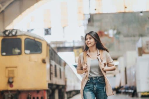 A woman walks along a train platform, with a train in the background. She’s carrying a backpack and looking up.