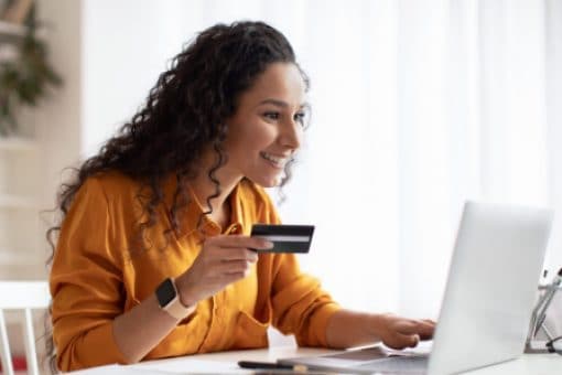 Smiling woman in orange shirt holds a credit card and types on her laptop.