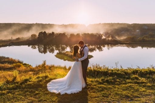 Bride and groom stand near a lake at sunset.