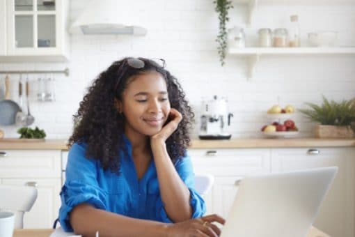 Smiling woman uses laptop at kitchen table covered with papers, a pen, and a coffee cup.