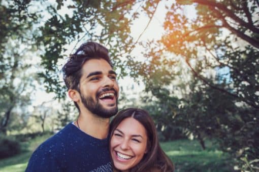 A happy couple smiles and embraces in a wooded area.