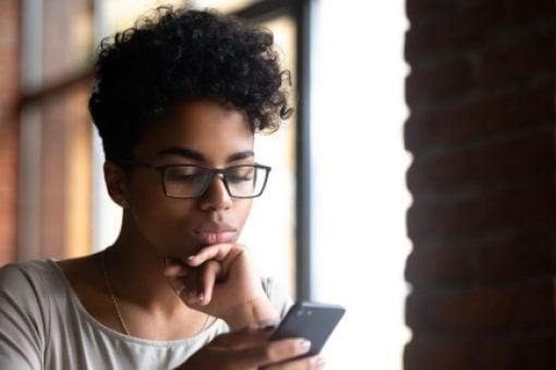 Woman with glasses sitting by window stares pensively at cell phone in hand
