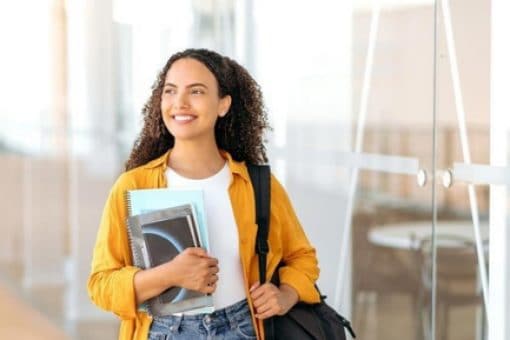 A student carrying notebooks and wearing a yellow shirt stands in a modern building with glass doors.