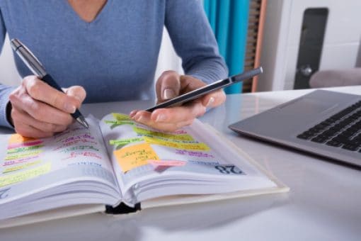 A person writes in a planner while referring to their phone.