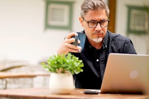 A man holds a coffee cup while he browses on his laptop computer.