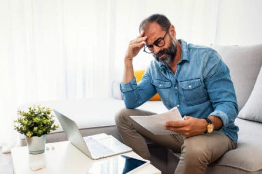 A man, wearing a blue button-up shirt and khaki pants, sits on a couch and stares at a document in his hands.