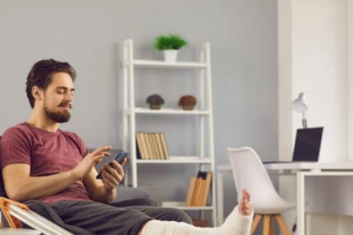 Man with a broken leg and crutches sits in his chair looking at a mobile phone.