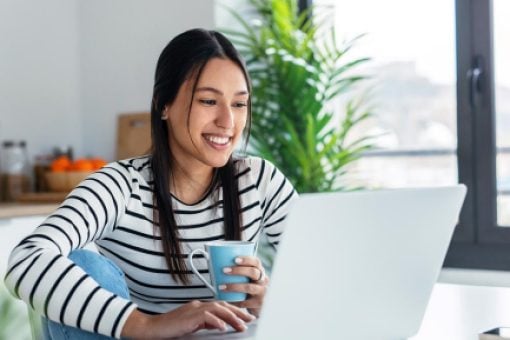Happy young woman sitting at her table, focused on her laptop, and enjoying a cup of coffee.
