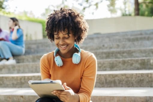 Woman with headphones around her neck, sitting on outdoor steps, engrossed in her tablet.