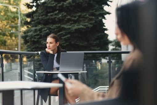 Woman sits outside at a table looking away from her laptop.