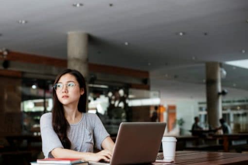 Young woman sits alone at cafeteria table with laptop and coffee.