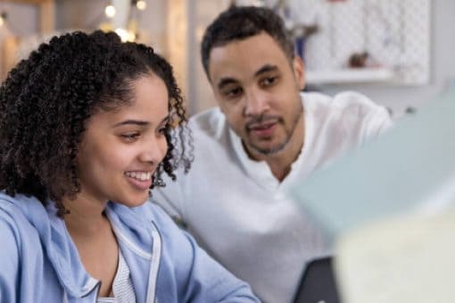 A young woman in a brightly lit room looks at a laptop with her father.