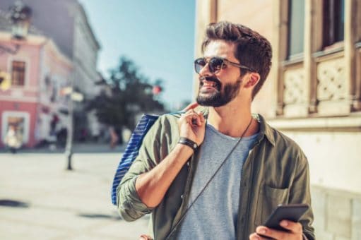 A smiling man walks down a street while traveling holding a shopping bag and a cell phone.