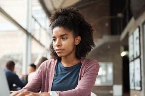 Young woman uses a laptop at a sunlit table with an open book beside her in a spacious office setting.