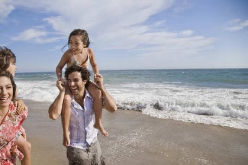 A happy family vacationing at the beach by the waves with kids resting on their parents' shoulders.