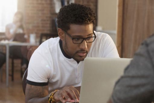 Young man with glasses looks at his laptop in a co-working space with people working at a desk in the background.