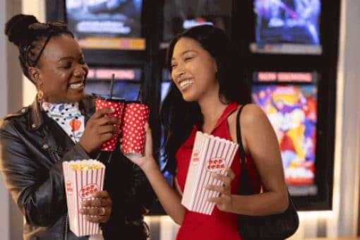 Two women with popcorn and drinks in a movie theater lobby.