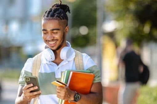 Smiling college student uses his smartphone on a sunny campus walkway.