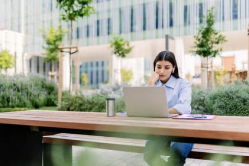 A woman sits at a outdoor table wearing earbuds and looking at a laptop. 
