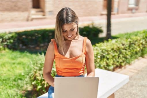 Woman uses laptop sitting outside in the sun on sidewalk bench