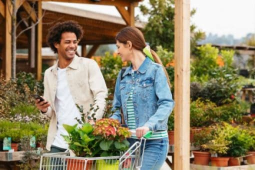 Two people shopping for plants together at a garden center, pushing a cart filled with greenery.