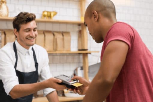 Barista handing a card reader to a customer making a payment with a credit card at a coffee shop counter.