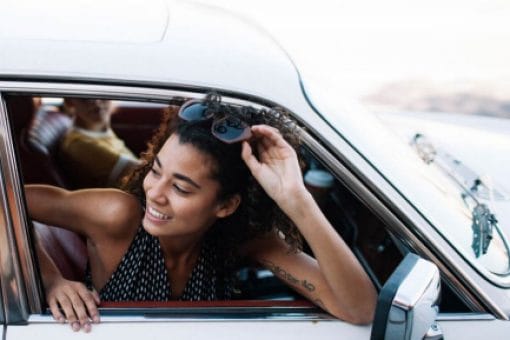 A smiling woman leans out of the car passenger window on a sunny day.