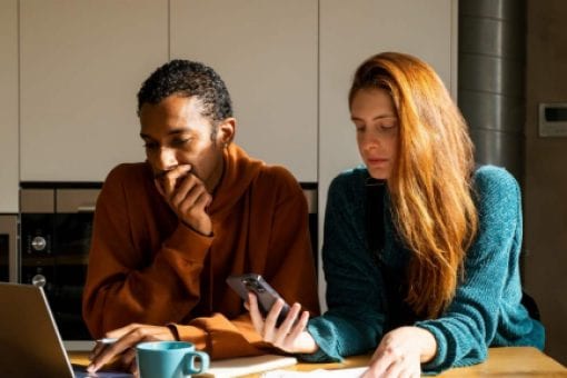 A couple sit together at a table looking thoughtfully at a laptop, phone, and paperwork.