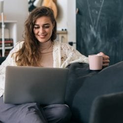 Smiling woman sitting on a couch with a laptop and holding a coffee mug in a cozy living room.