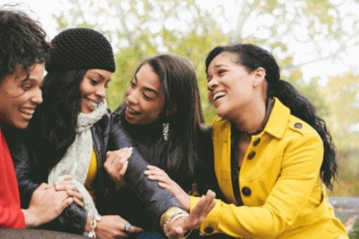 Three women look on excitedly as a fourth woman holds her left hand out showing her engagement ring.