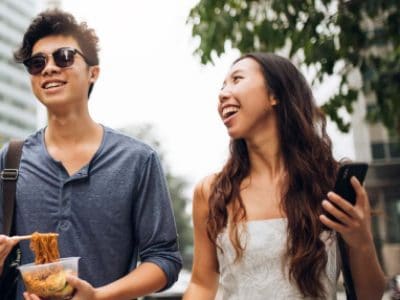 Two friends walking outside and smiling, one holding a phone and one eating.