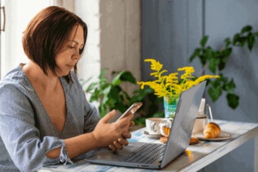 A woman sits at a table with a laptop and phone, and breakfast items and flowers on the table.