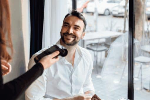 Man in a white shirt sits at a restaurant table ready to make a payment with a credit card while a server holds a payment terminal.