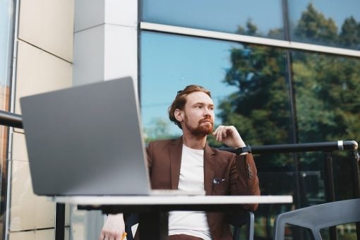Man sits outside at a table looking away from his laptop with large, glass office building behind him.