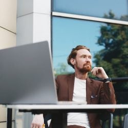 Man sits outside at a table looking away from his laptop with large, glass office building behind him.