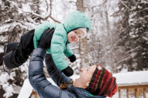 Mother holding up a smiling child in a snowy outdoor setting.