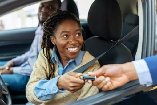A person handing car keys to a couple who are sitting in a car looking happy.