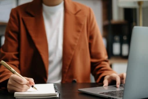 Person sits in front of laptop and writes in a notebook.