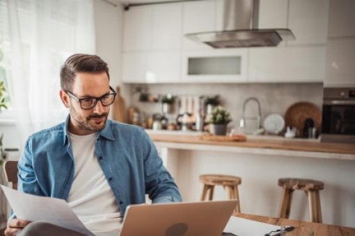 Person using a laptop and reviewing documents at a kitchen table.