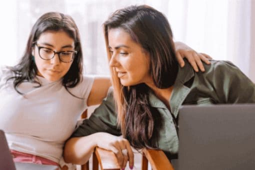 Two women sit together, looking at laptop screens, with one placing her arm around the other’s shoulders.