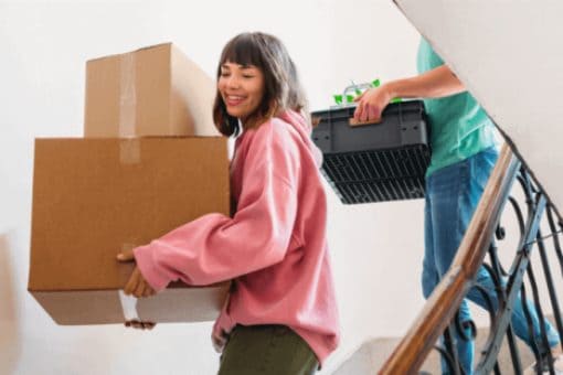 Woman in a pink sweater carrying boxes down a stairwell followed by a man carrying a plastic container.