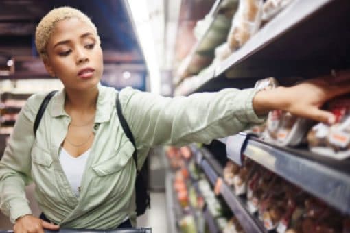 A woman pushing a cart through a grocery store reaches onto a shelf for some produce.