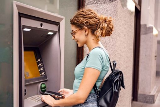 A woman stands at an ATM and holds a mobile phone.