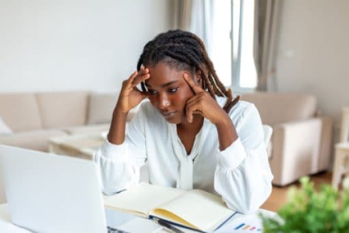 A young woman sits at a table comparing her laptop screen with a notebook.