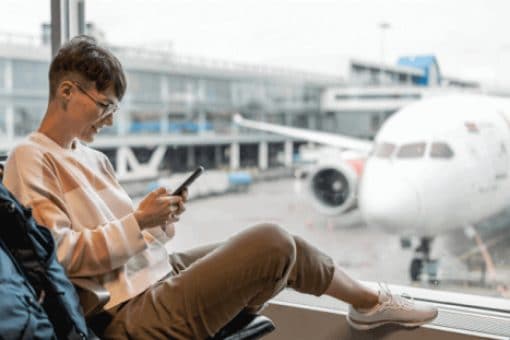Traveler sitting in an airport terminal by a window using a smartphone with a plane in the background.