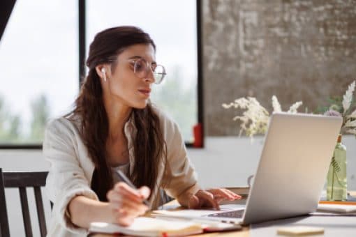 A Young Lady with glasses and headphones sits at a table looking at a laptop and writing in a notebook.
