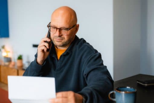 A man sitting at a kitchen counter holds a phone to his ear while holding a paper document in his other hand.