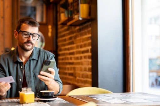 Man sitting in a café holds a credit card in one hand and looks at his smartphone in his other hand, with coffee and orange juice on the table.